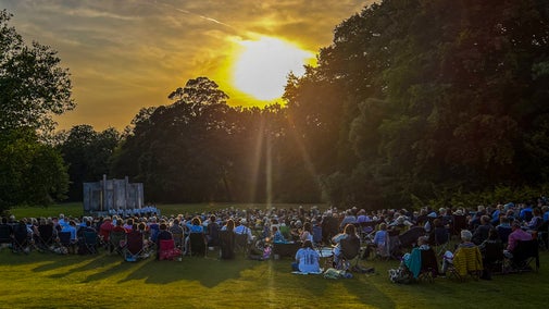 An audience watching an outdoor theatre performance in a parkland at sunset